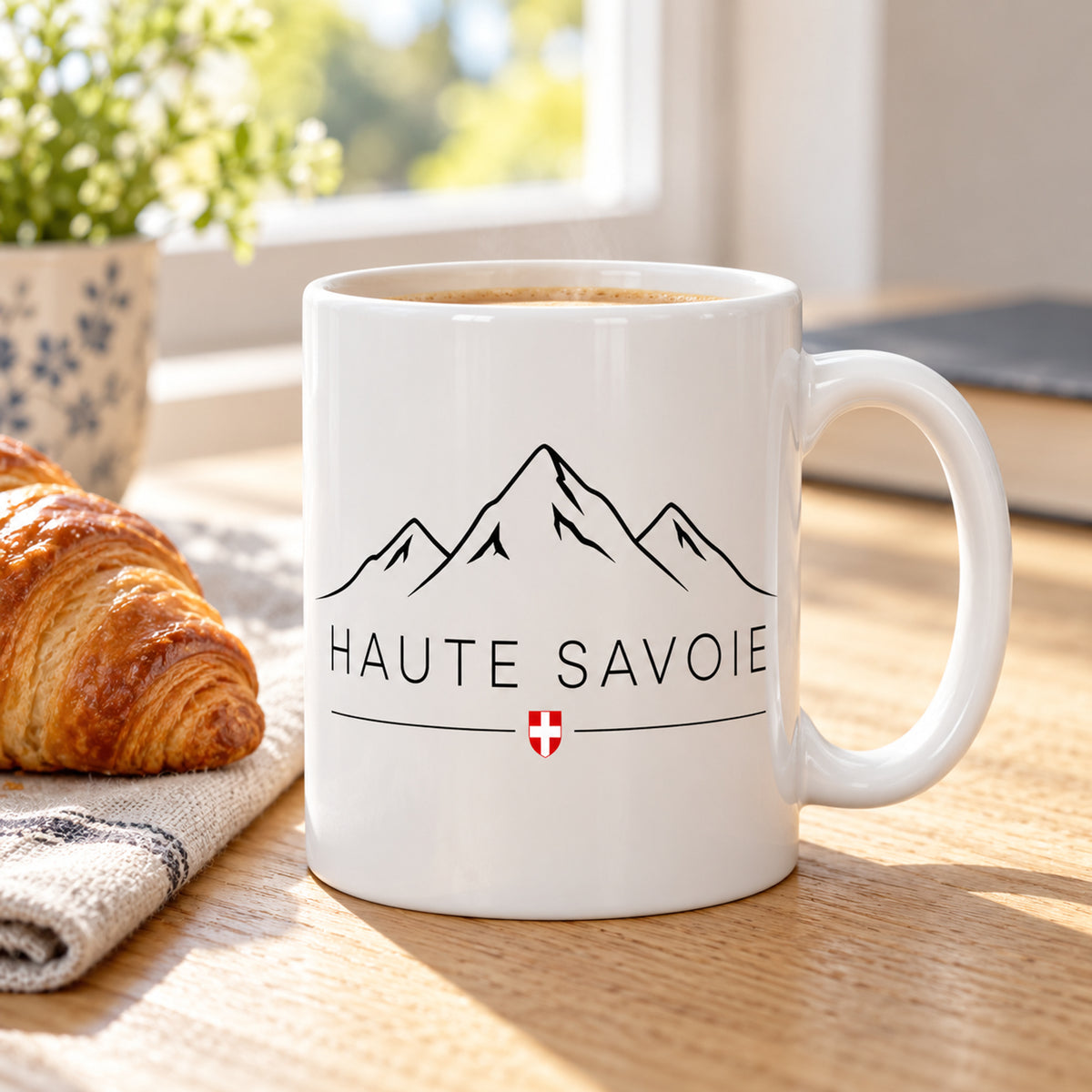 Mug Haute-Savoie « Sommets & Écusson Savoyard » posé sur une table en bois avec un croissant, pause café