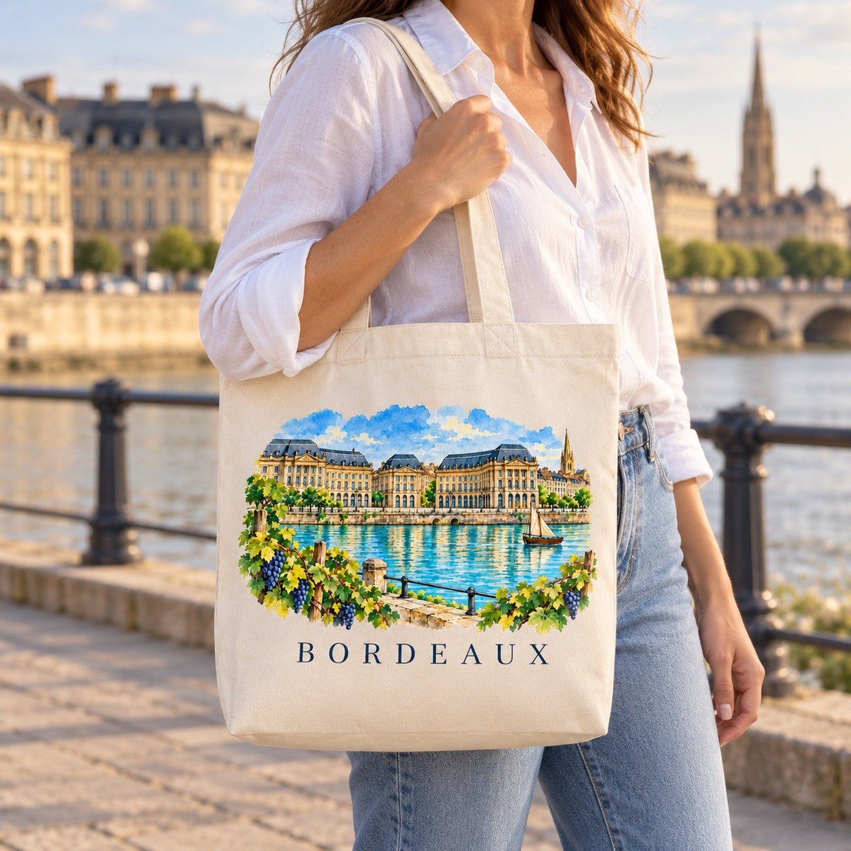Tote bag Bordeaux Premium porté à l'épaule sur les quais de la Garonne, façades et pont de pierre en arrière-plan
