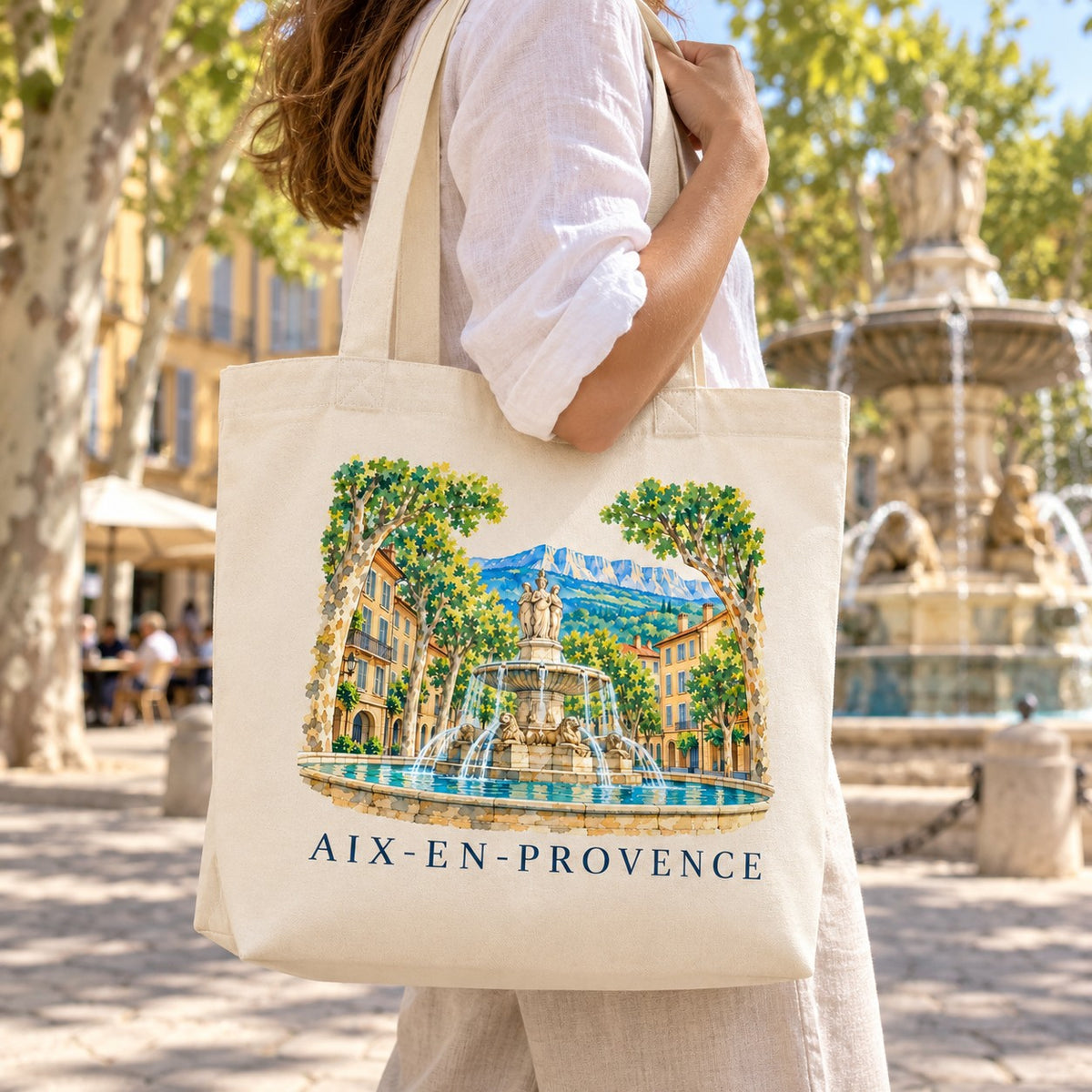 Tote bag Aix-en-Provence Premium porté à l'épaule près d'une fontaine sous les platanes