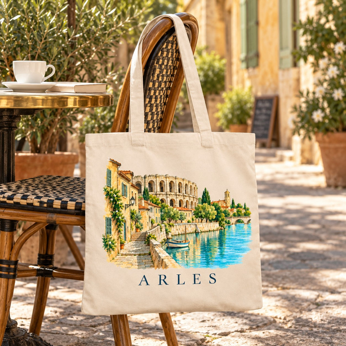 Tote bag Arles Essentiel suspendu à une chaise en terrasse dans une ruelle ensoleillée, avec un café
