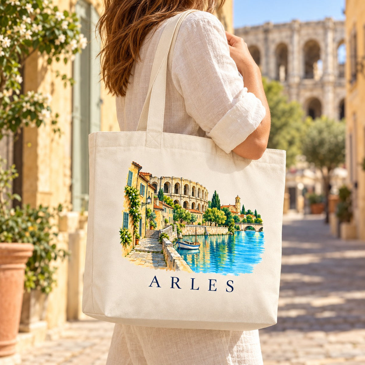 Tote bag Arles Premium porté à l'épaule dans une ruelle ocre, arènes en arrière-plan