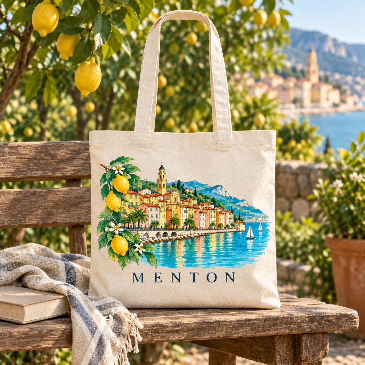Tote bag Menton Essentiel posé sur un banc sous un citronnier, avec un livre et un plaid