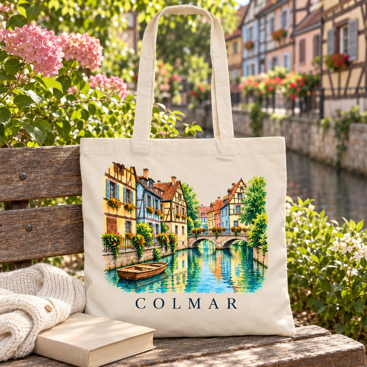 Tote bag Colmar Essentiel posé sur un banc fleuri au bord de l'eau, avec un livre et un pull