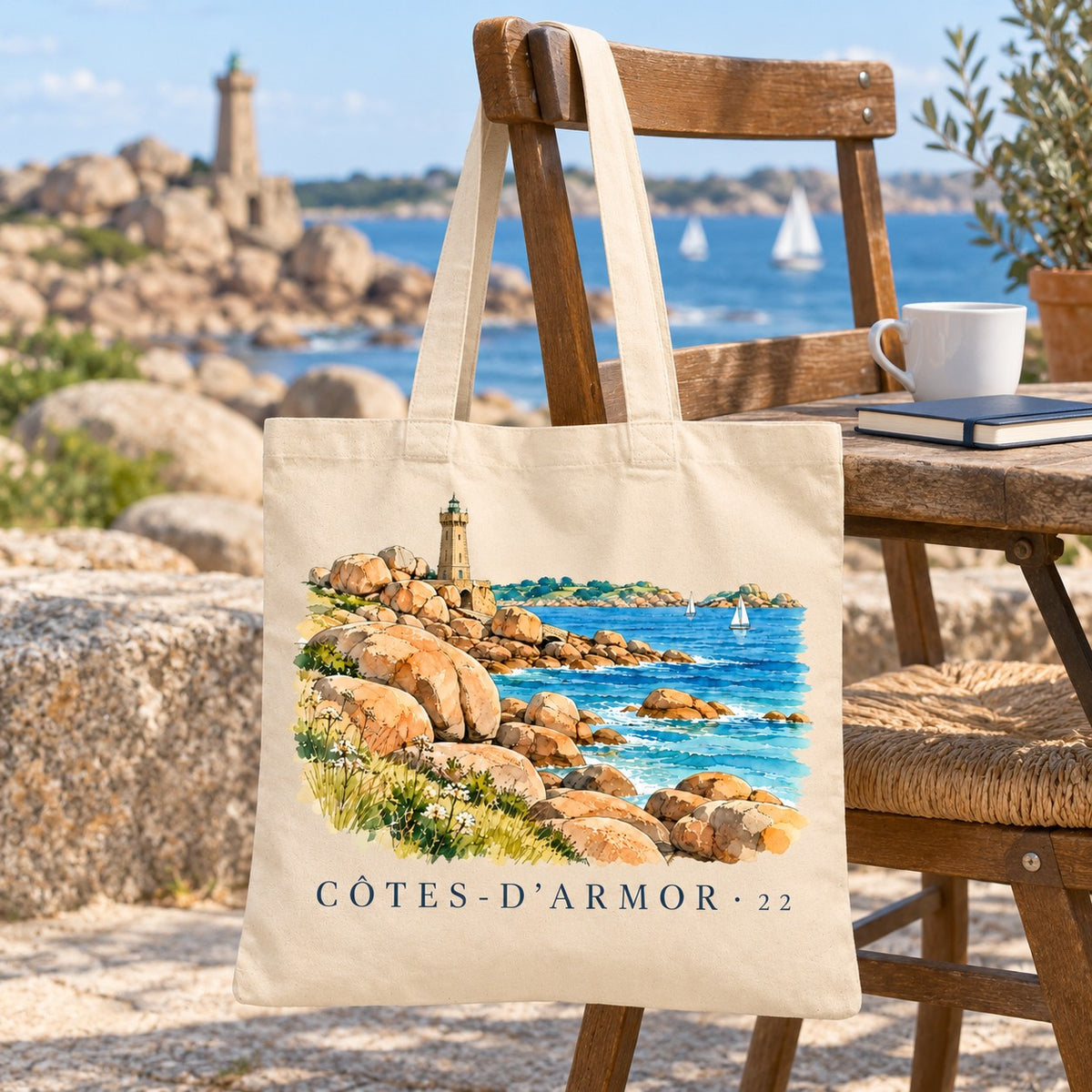 Tote bag Côtes-d'Armor (22) Essentiel suspendu à une chaise en terrasse face aux rochers de granit rose et au phare, avec un café