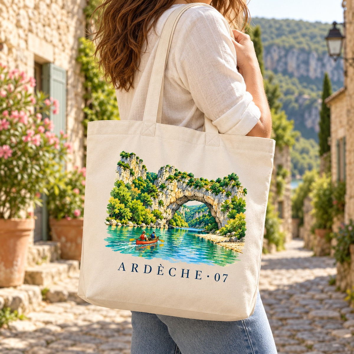Tote bag Ardèche (07) Premium porté à l'épaule dans une ruelle de village en pierre, gorges en arrière-plan