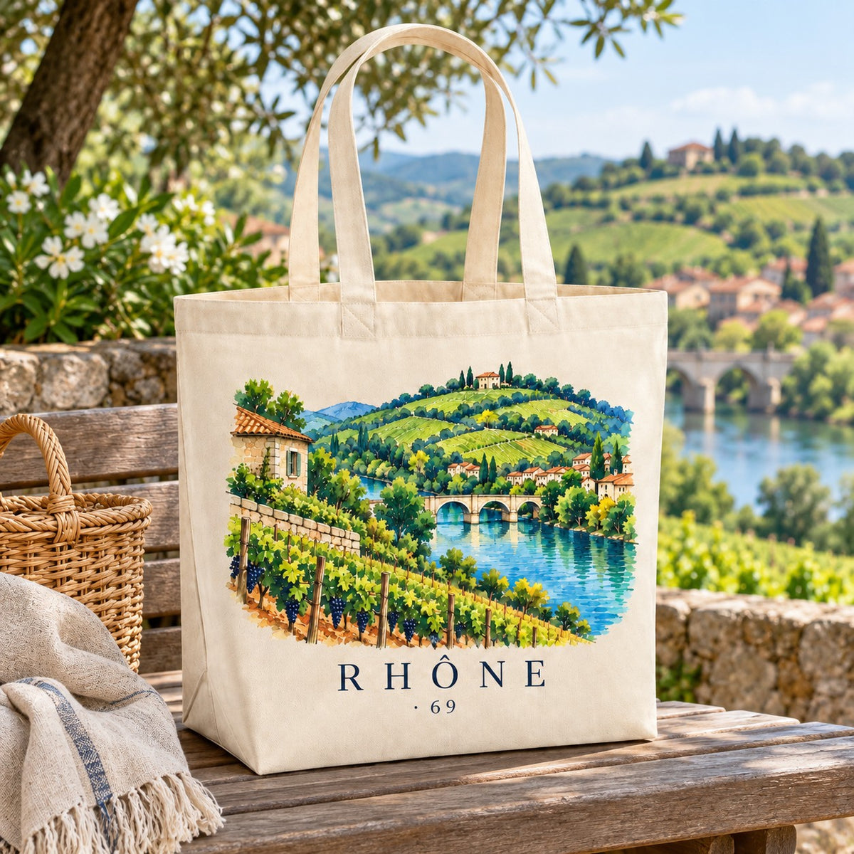 Tote bag Rhône (69) Premium posé sur un banc de bois, avec un panier et un plaid, pont de pierre en arrière-plan