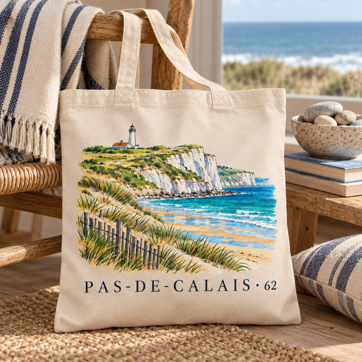 Tote bag Pas-de-Calais (62) Essentiel posé contre un fauteuil en rotin près d'une fenêtre ouverte sur la mer