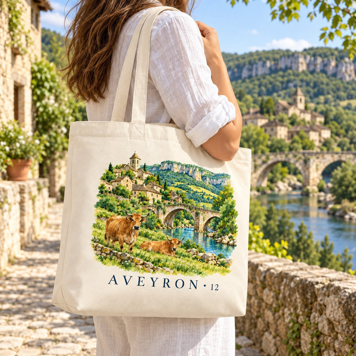 Tote bag Aveyron (12) Premium porté à l'épaule dans une ruelle de pierre, pont et village en arrière-plan