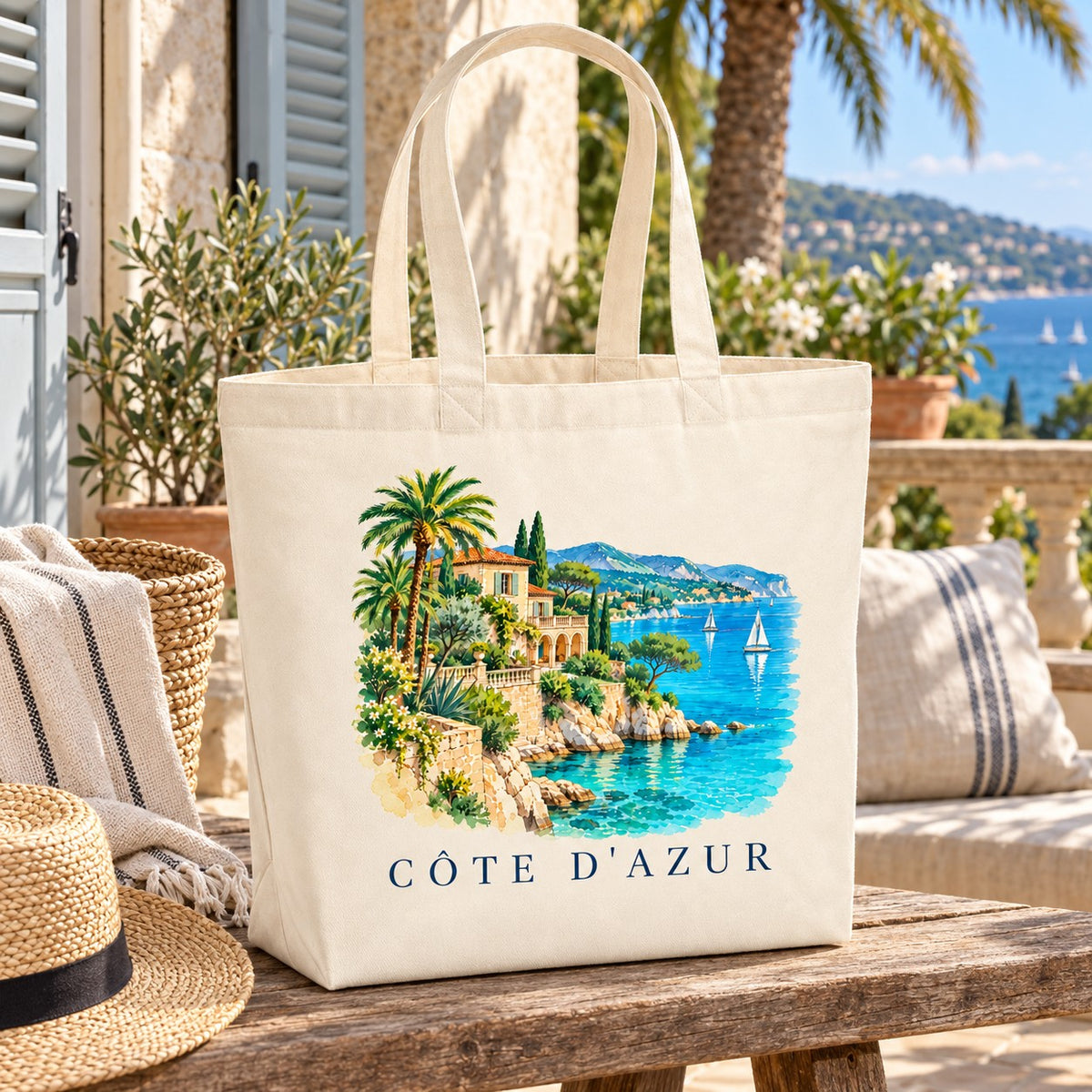 Tote bag Côte d'Azur Premium posé sur une table en terrasse face à la mer, avec un panier et un chapeau de paille