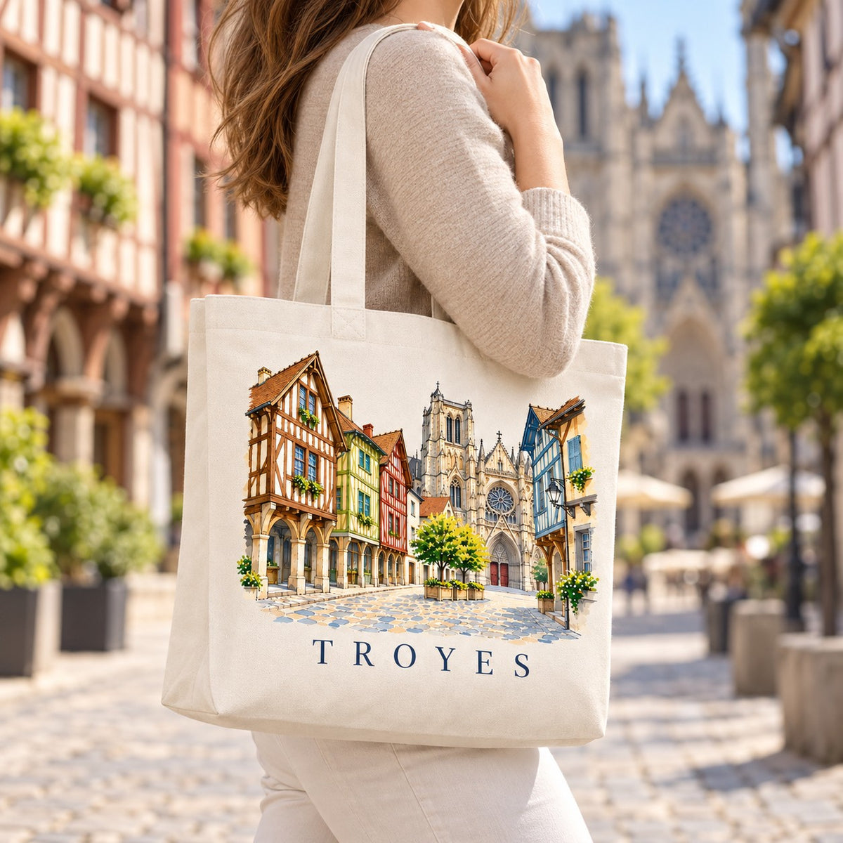 Tote bag Troyes Premium porté à l'épaule dans une rue pavée bordée de maisons à pans de bois, cathédrale en arrière-plan