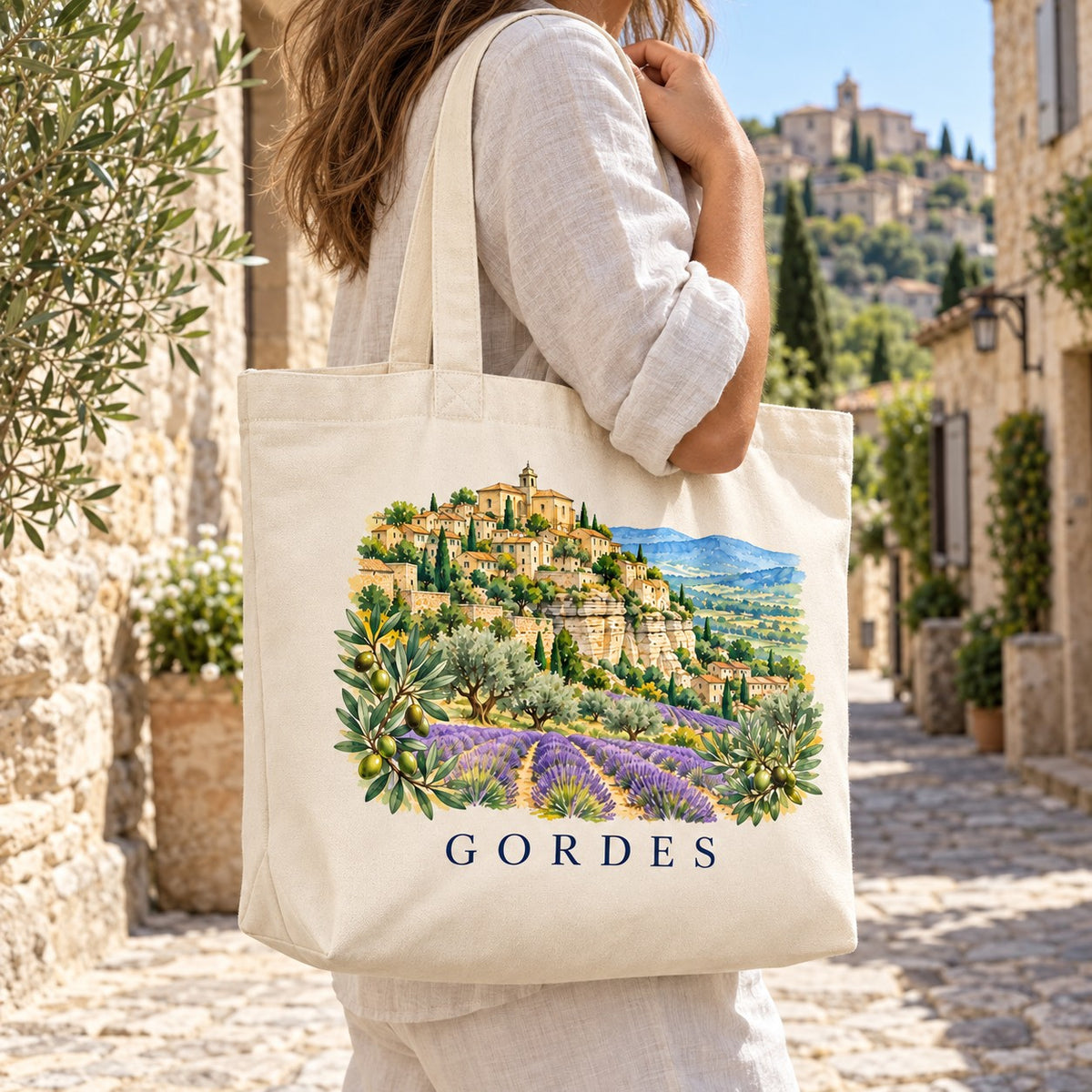 Tote bag Gordes Premium porté à l'épaule dans une ruelle de pierre, village perché en arrière-plan
