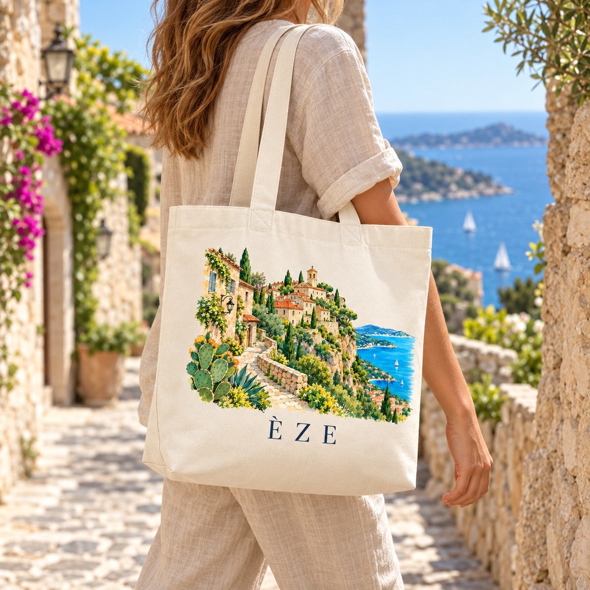 Tote bag Èze Premium porté à l'épaule dans une ruelle de pierre fleurie de bougainvilliers, mer en arrière-plan