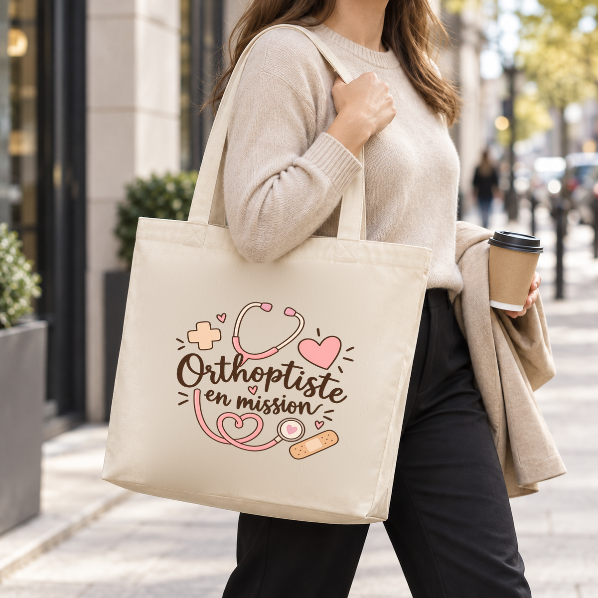 Tote bag orthoptiste « Orthoptiste en mission » Premium porté à l'épaule dans la rue, un café à la main