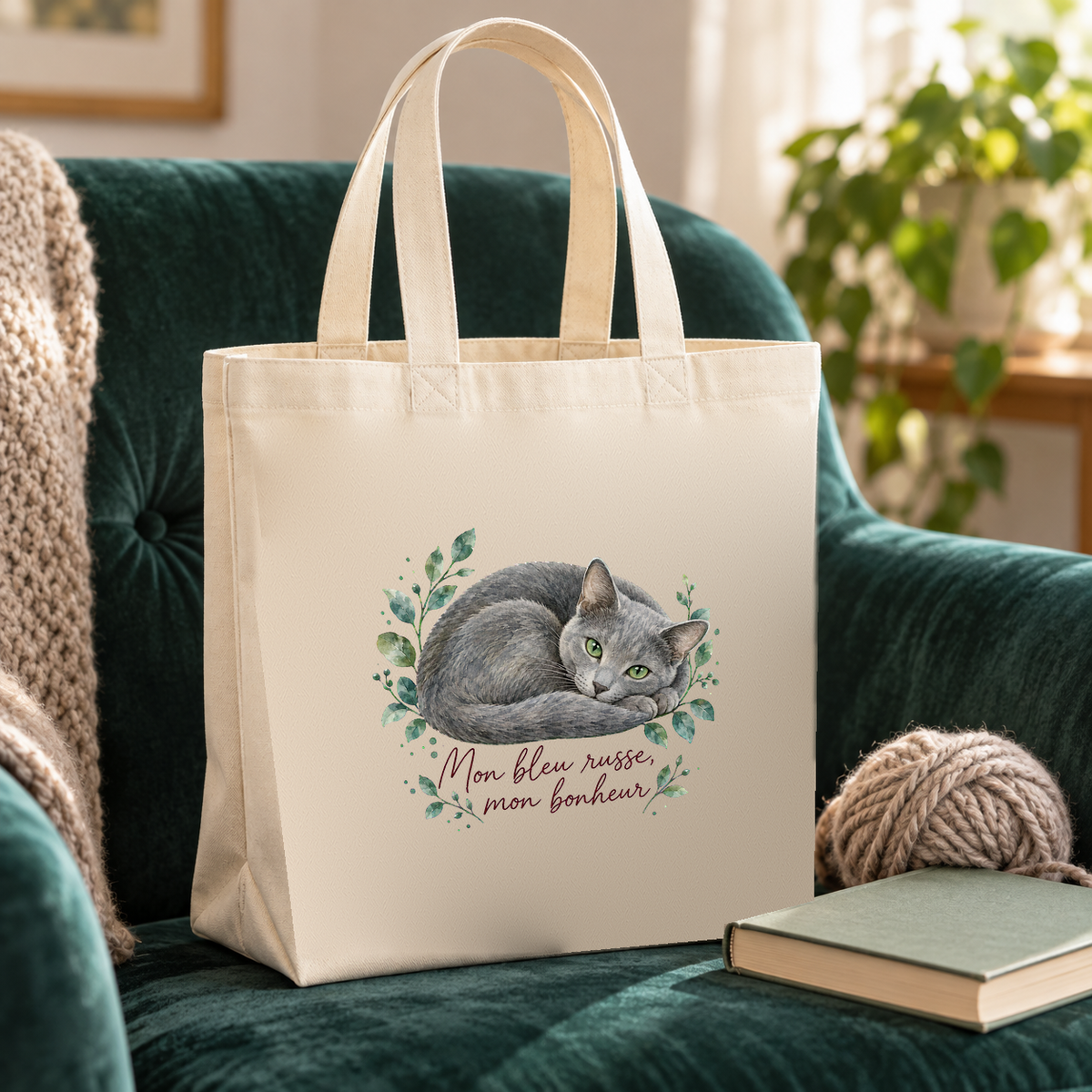 Tote bag bleu russe « Mon bleu russe, mon bonheur » Premium posé sur un fauteuil en velours vert émeraude, à côté d'une pelote de laine et d'un livre