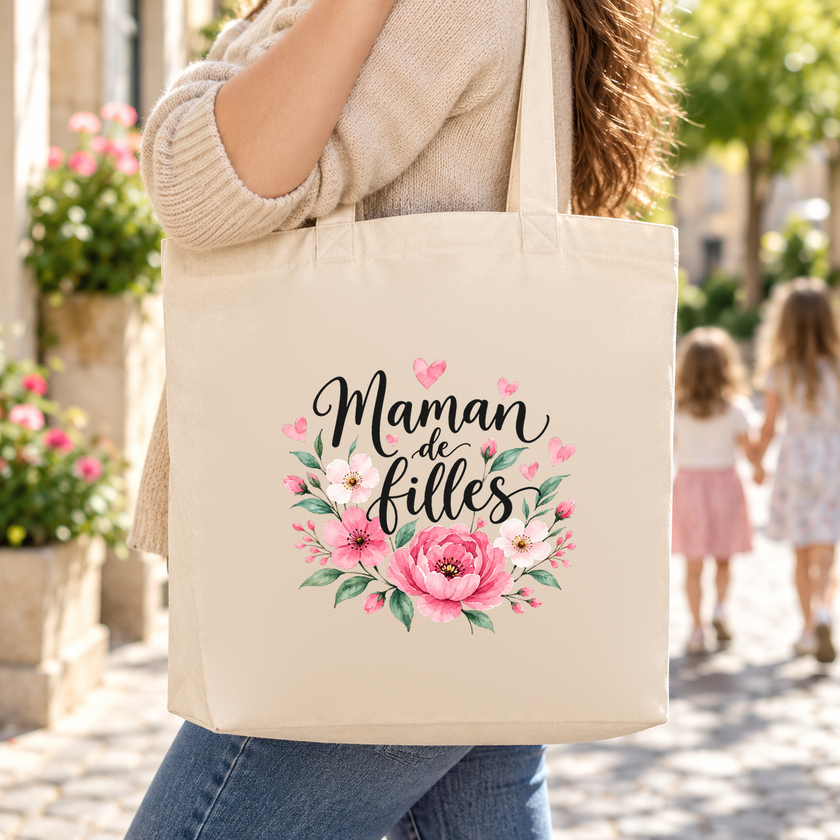 Tote bag maman « Maman de filles » Premium porté à l'épaule dans une rue fleurie, deux petites filles main dans la main