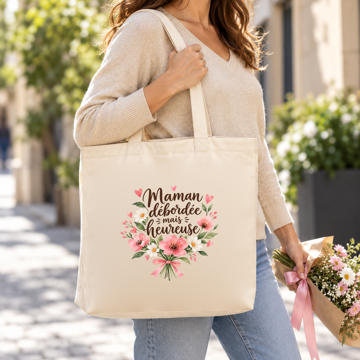 Tote bag maman « Maman débordée mais heureuse » Premium porté à l'épaule dans une rue ensoleillée, un bouquet à la main