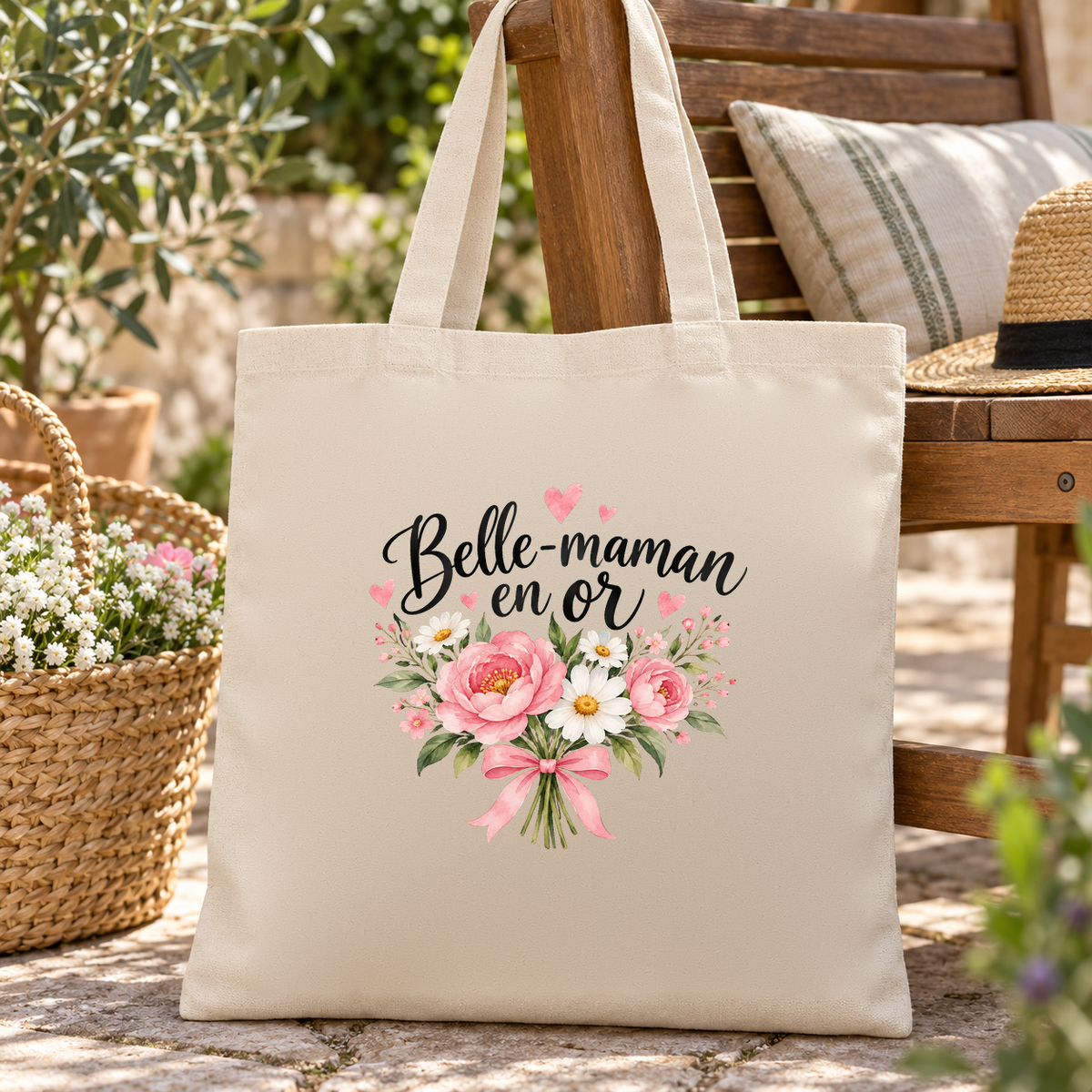 Tote bag belle-maman « Belle-maman en or » Essentiel posé contre une chaise de jardin en bois, à côté d'un panier de fleurs et d'un chapeau de paille