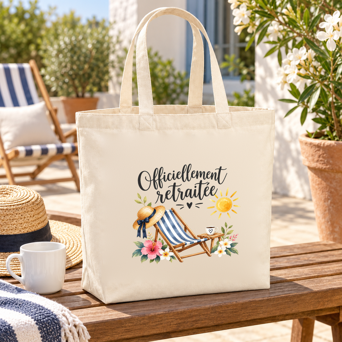 Tote bag retraite « Officiellement retraitée » Premium posé sur une table de terrasse, à côté d'un chapeau de paille et d'une tasse de café