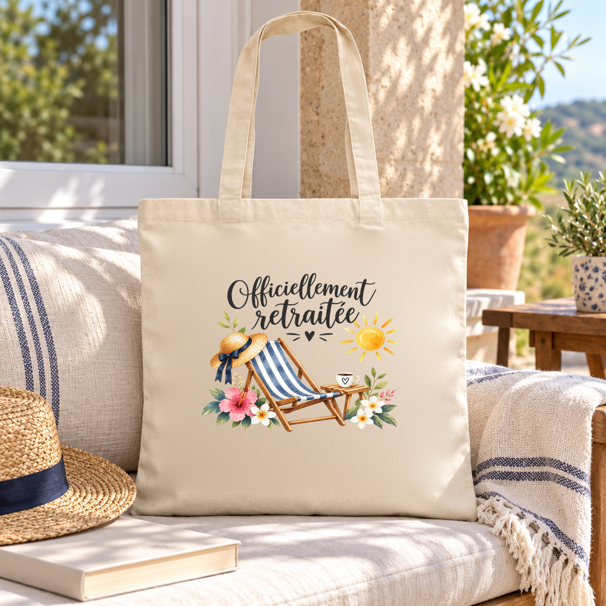 Tote bag retraite « Officiellement retraitée » Essentiel posé sur une banquette de terrasse, à côté d'un chapeau de paille et d'un livre