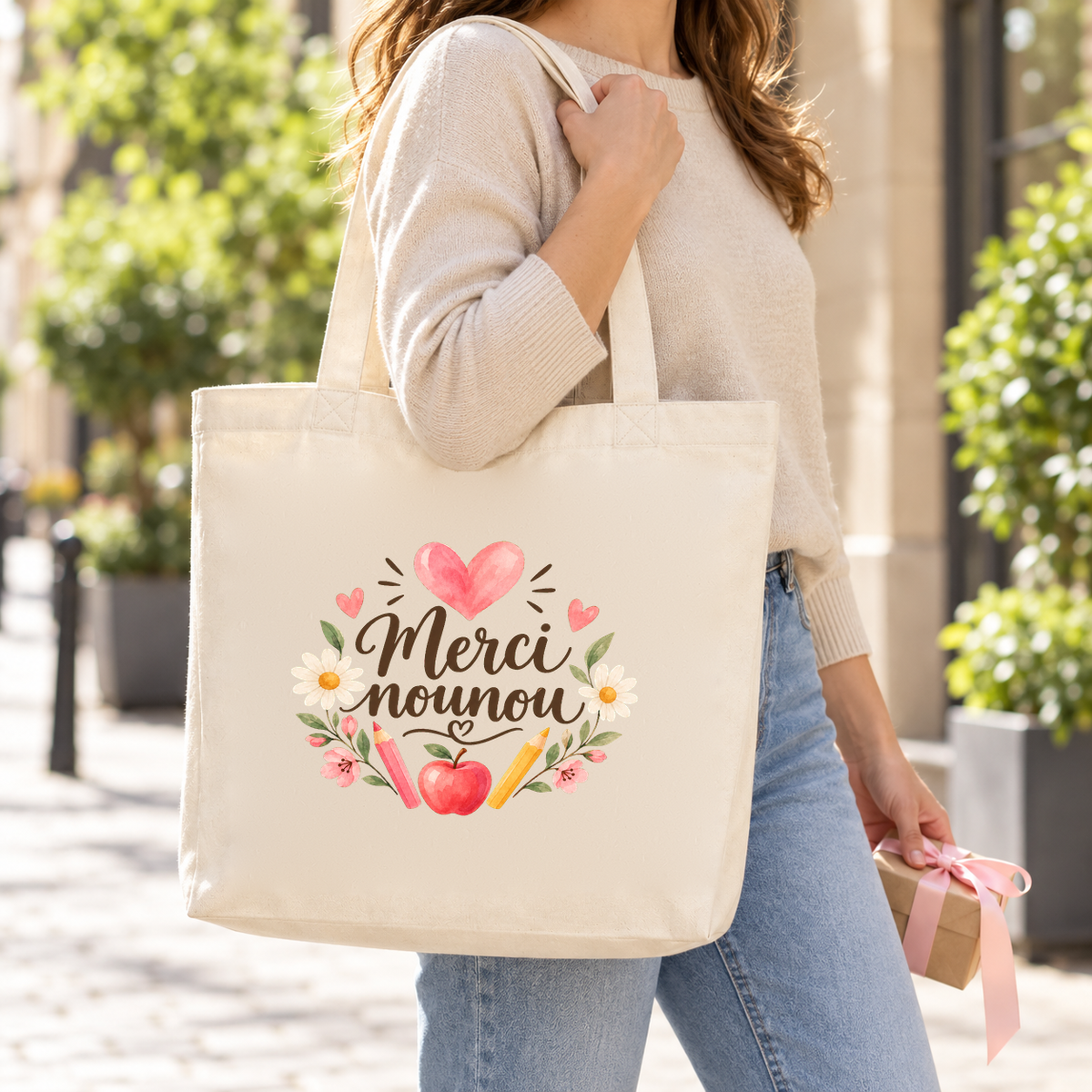 Tote bag nounou « Merci nounou » Premium porté à l'épaule dans une rue ensoleillée, un petit cadeau à la main