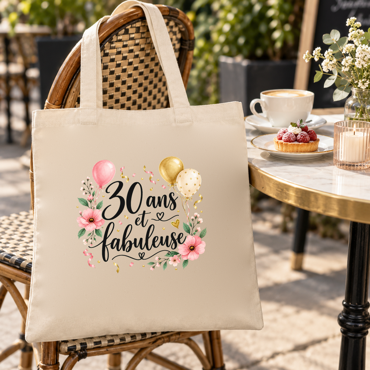 Tote bag anniversaire « 30 ans et fabuleuse » Essentiel suspendu à une chaise de terrasse en rotin, à côté d'un café et d'une tartelette aux framboises