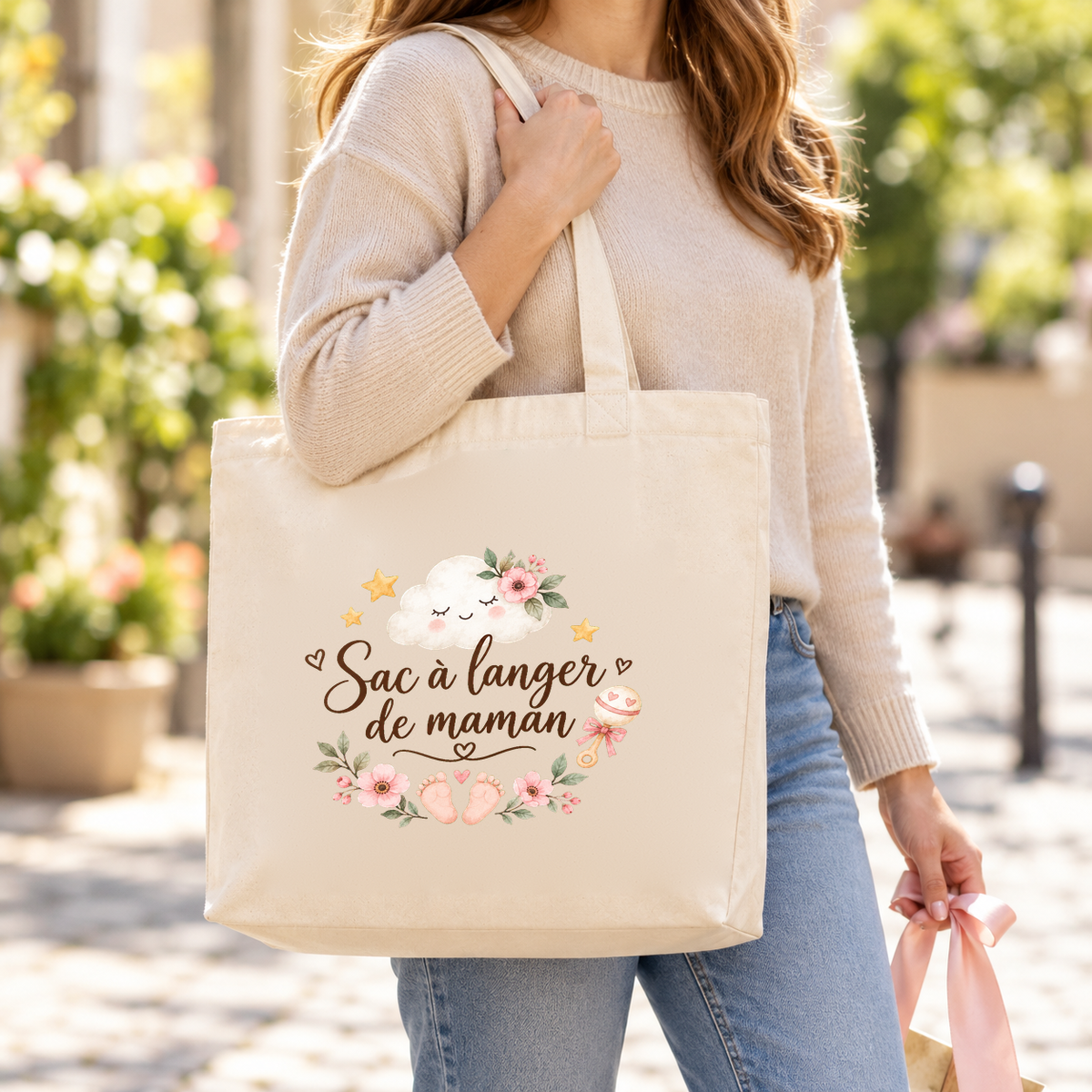 Tote bag naissance « Sac à langer de maman » Premium porté à l'épaule dans une rue ensoleillée, un petit cadeau à la main