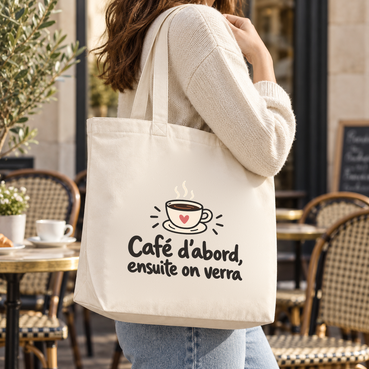 Tote bag « Café d'abord, ensuite on verra » Premium porté à l'épaule devant une terrasse de café