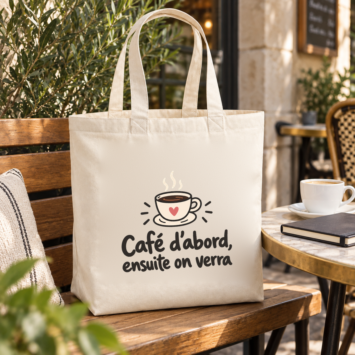 Tote bag « Café d'abord, ensuite on verra » Premium posé sur une banquette de terrasse, à côté d'un café et d'un carnet