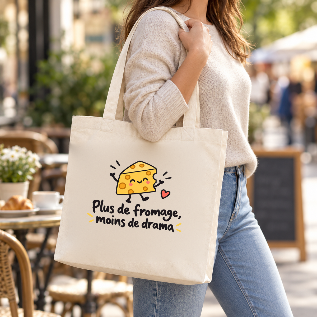 Tote bag « Plus de fromage, moins de drama » Premium porté à l'épaule dans une rue animée, devant une terrasse