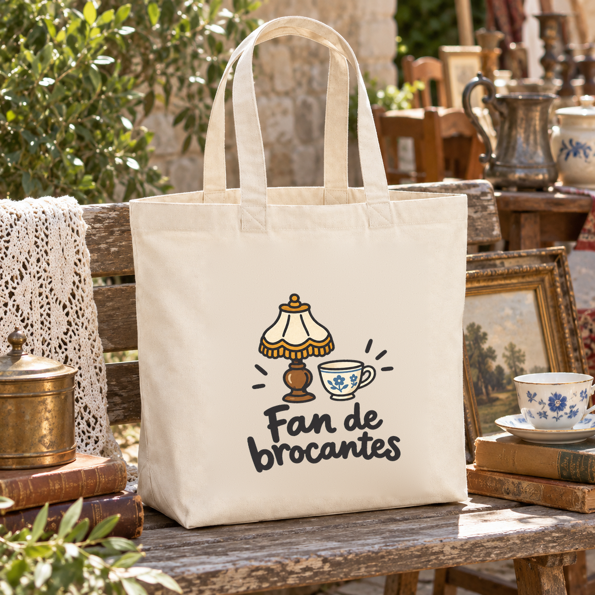 Tote bag « Fan de brocantes » Premium posé sur une table de brocante, entre cadres anciens, théière et vieux livres