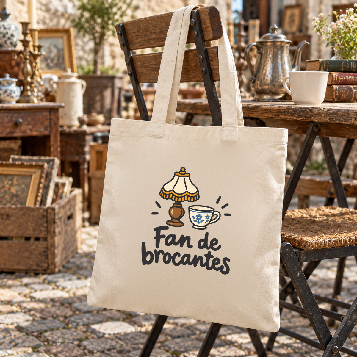 Tote bag « Fan de brocantes » Essentiel suspendu à une chaise en bois au milieu des stands d'une brocante