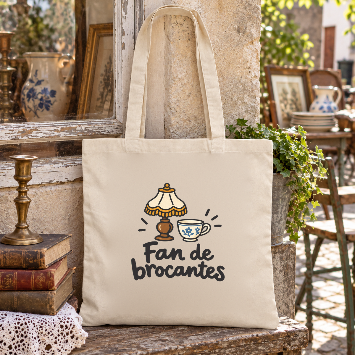 Tote bag « Fan de brocantes » Essentiel posé sur un banc en pierre, à côté de livres anciens, d'un chandelier et d'un pichet en faïence