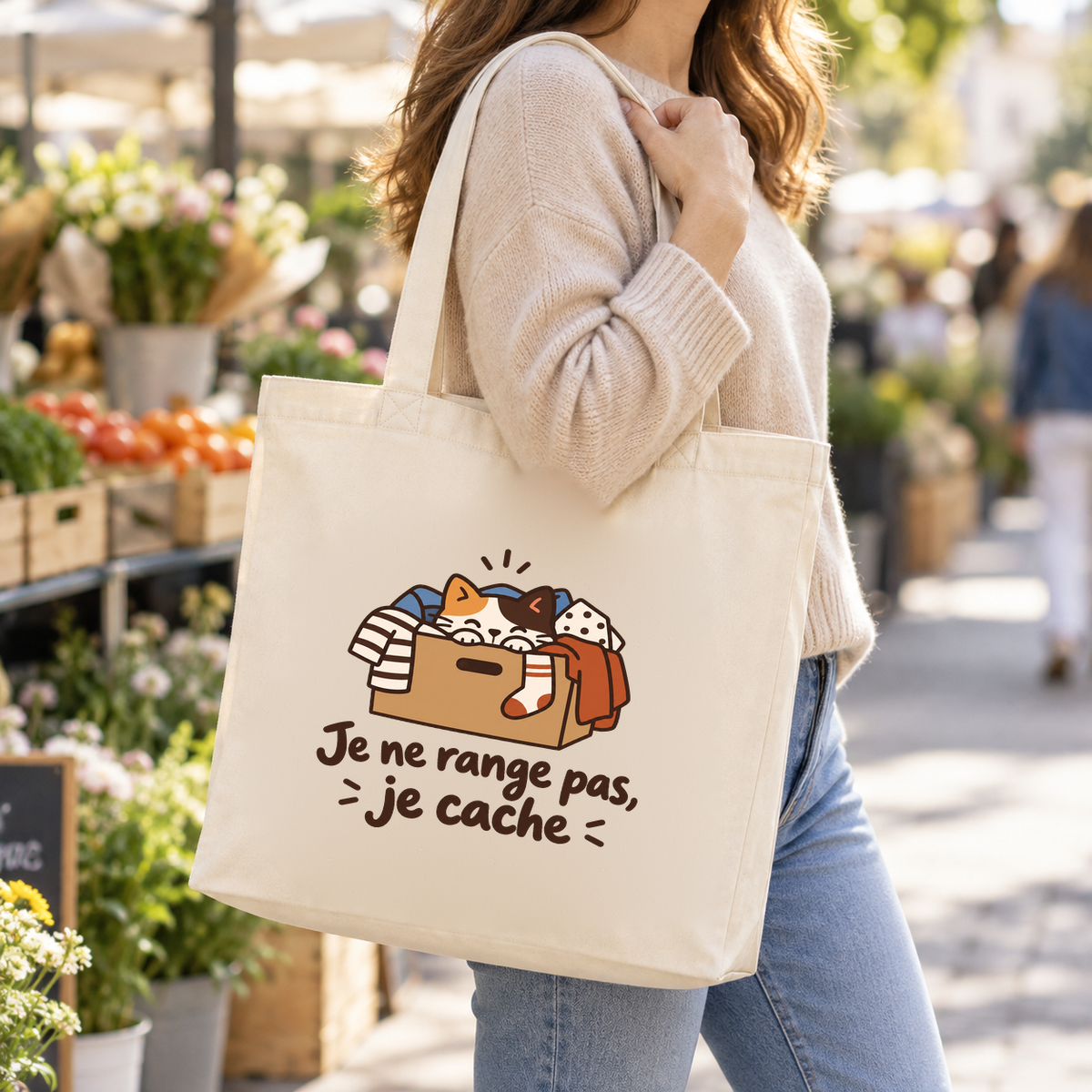 Tote bag « Je ne range pas, je cache » Premium porté à l'épaule dans une rue de marché
