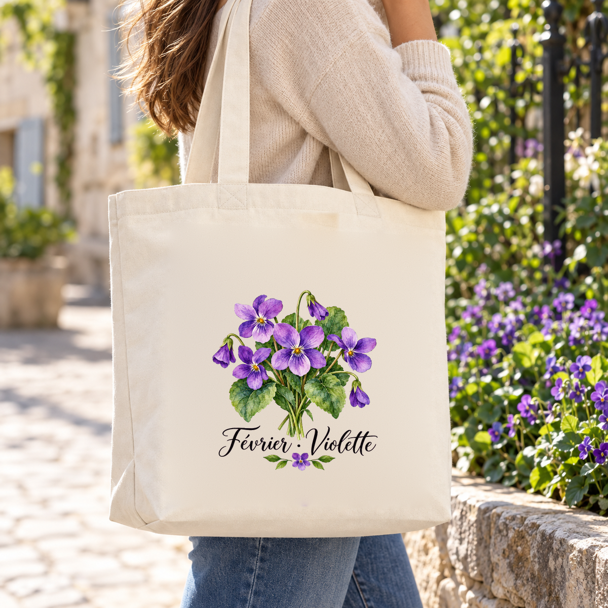 Tote bag fleur de naissance « Février · Violette » Premium porté à l'épaule dans une ruelle pavée fleurie de violettes