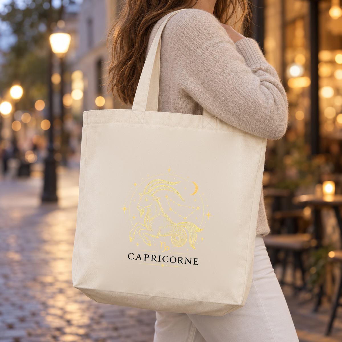 Tote bag signe astrologique « CAPRICORNE » Premium porté à l'épaule dans une rue pavée illuminée au crépuscule