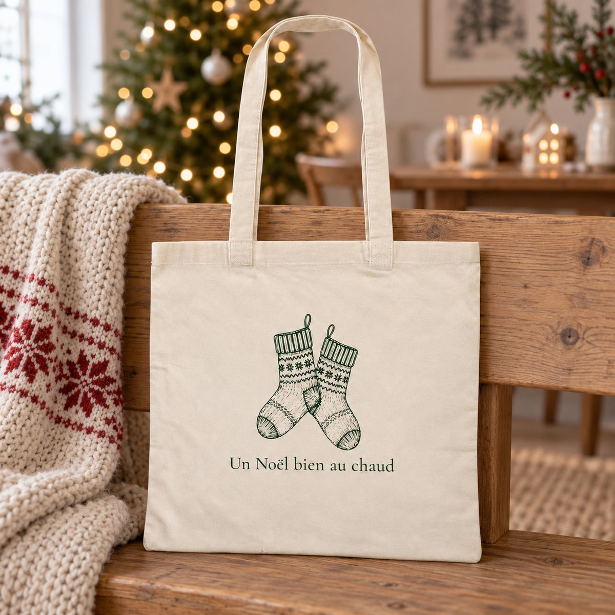 Tote bag de Noël « Un Noël bien au chaud » Essentiel appuyé contre un banc près d'un plaid de Noël