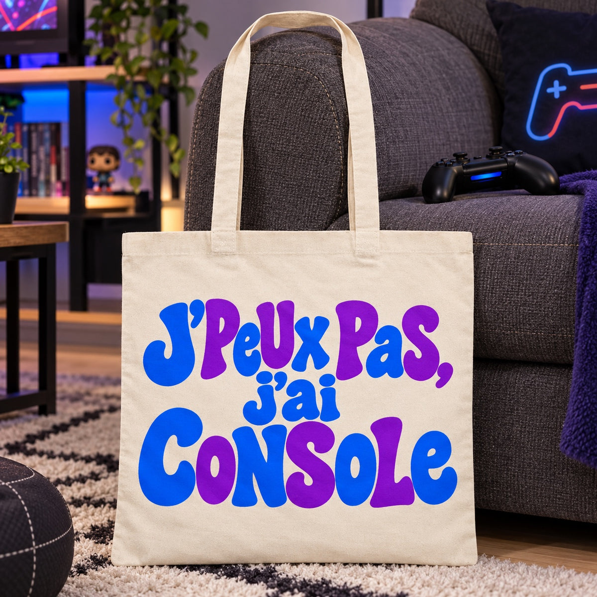 Tote bag « J'peux pas, j'ai console » Essentiel appuyé contre un canapé, manette posée sur l'accoudoir