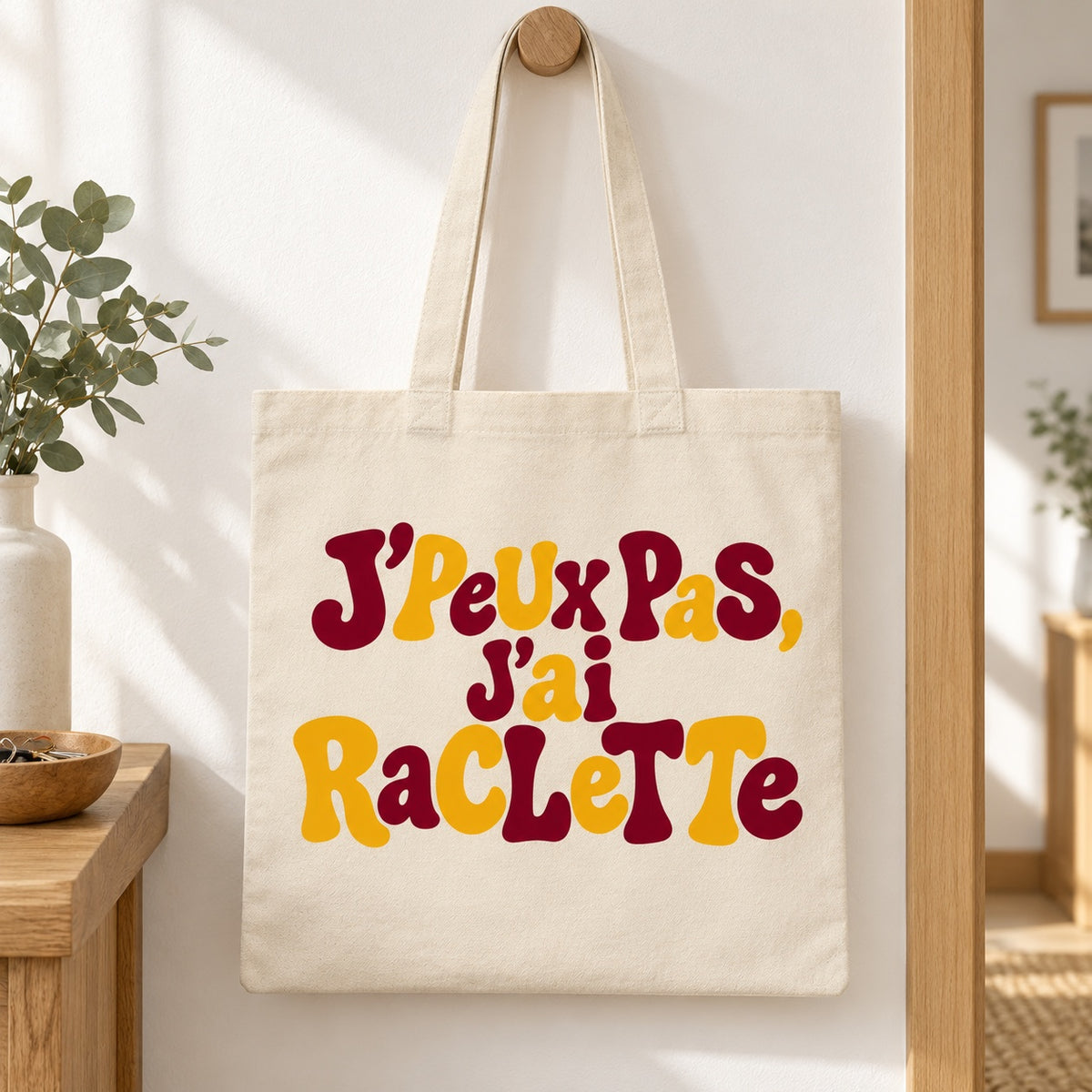 Tote bag « J'peux pas, j'ai raclette » Essentiel suspendu à une patère dans une entrée