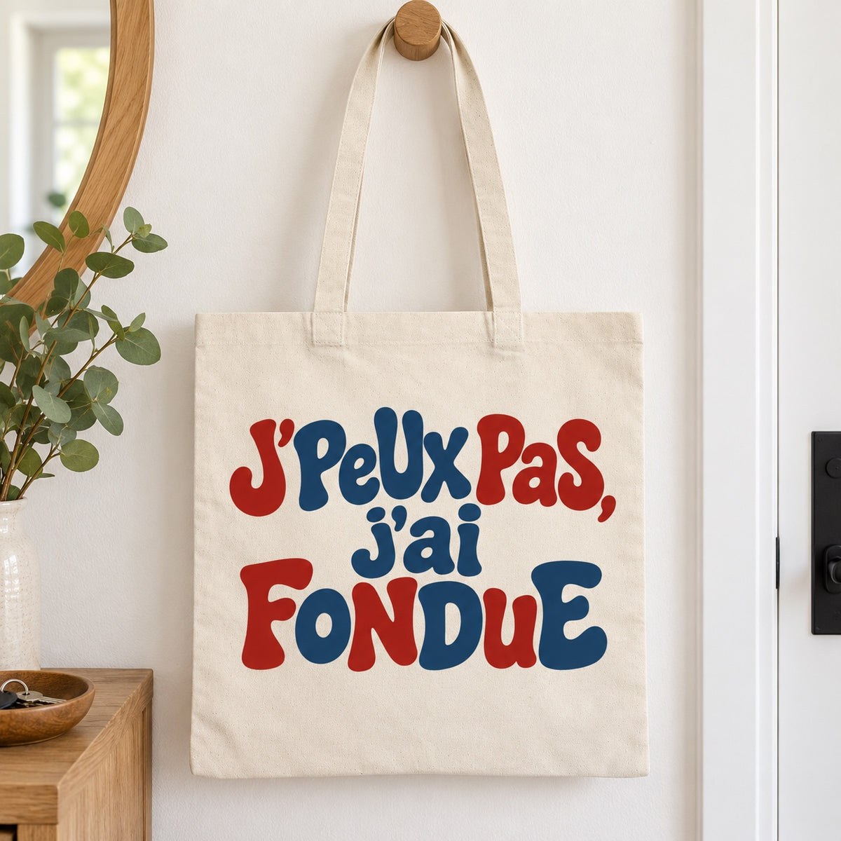 Tote bag « J'peux pas, j'ai fondue » Essentiel suspendu à une patère dans une entrée
