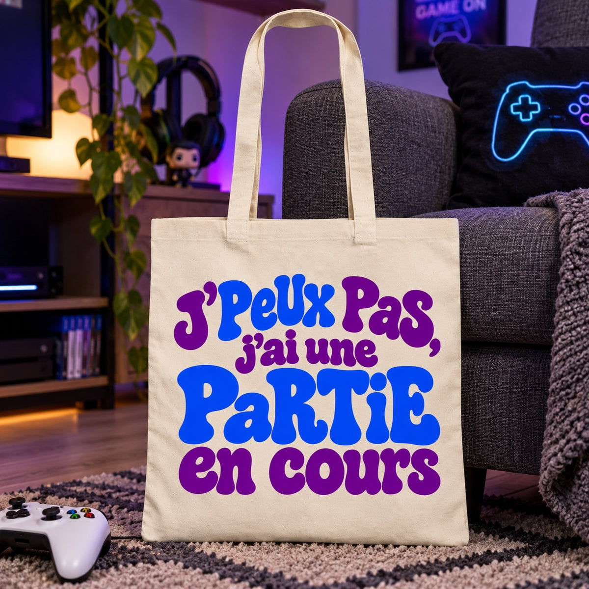 Tote bag « J'peux pas, j'ai une partie en cours » Essentiel appuyé contre un canapé près d'une manette