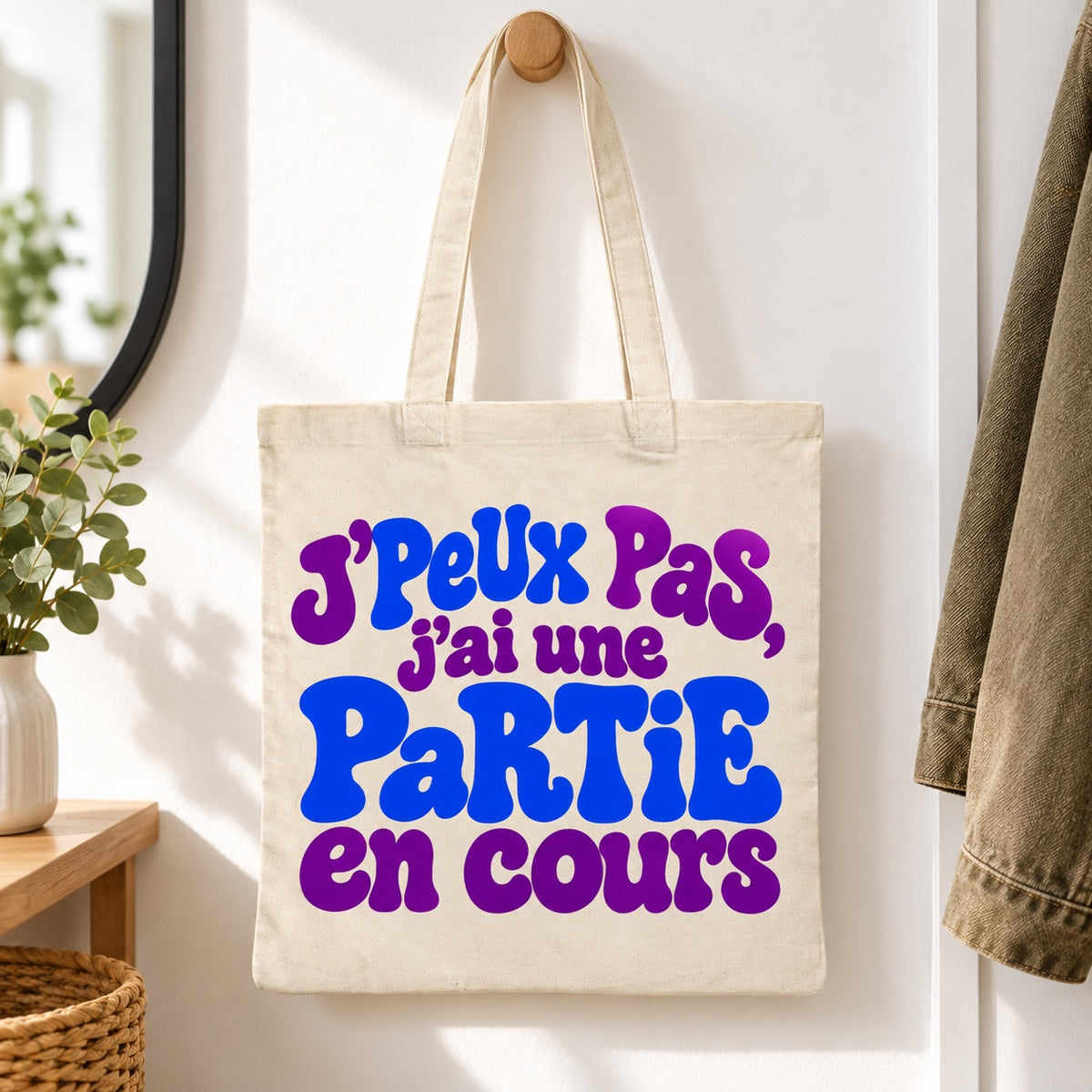 Tote bag « J'peux pas, j'ai une partie en cours » Essentiel suspendu à une patère dans une entrée