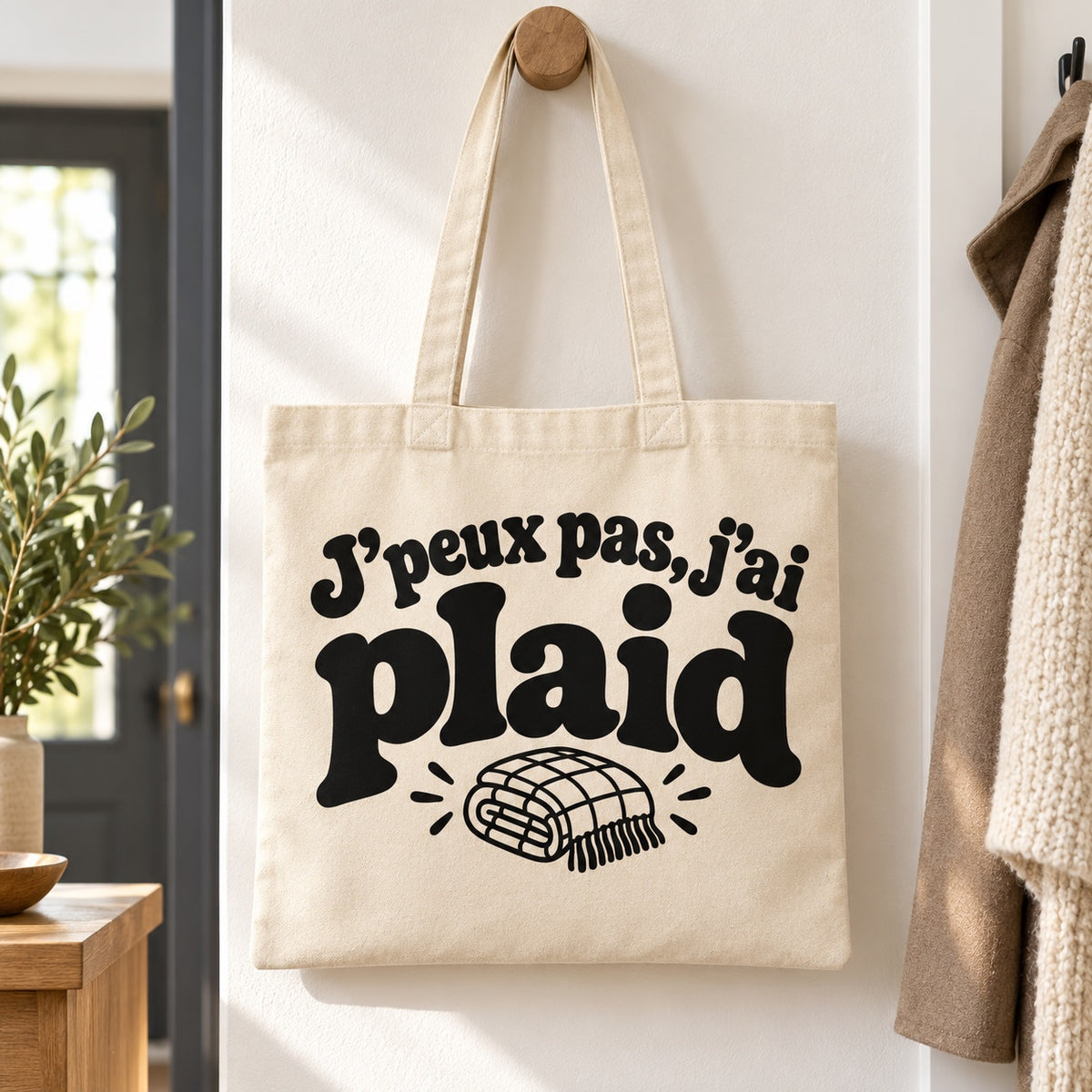 Tote bag « J'peux pas, j'ai plaid » Essentiel suspendu à une patère dans une entrée lumineuse