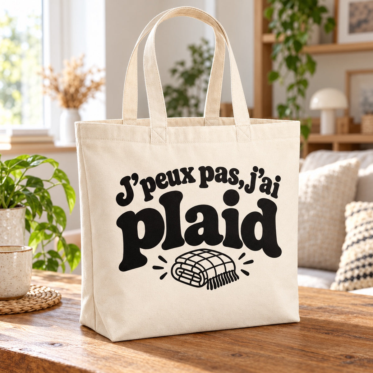 Tote bag « J'peux pas, j'ai plaid » Premium posé sur une table en bois dans un salon lumineux