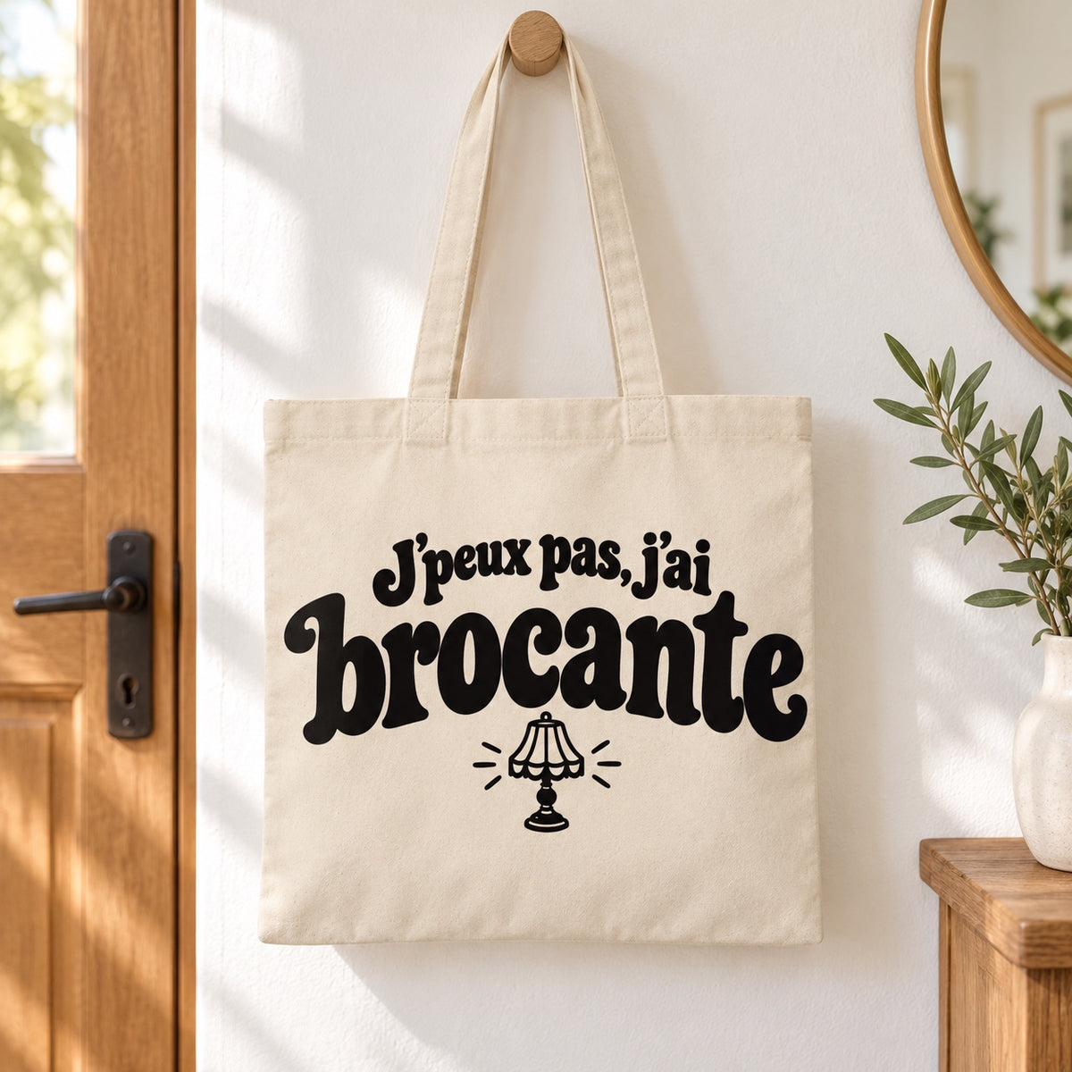 Tote bag « J'peux pas, j'ai brocante » Essentiel suspendu à une patère dans une entrée lumineuse
