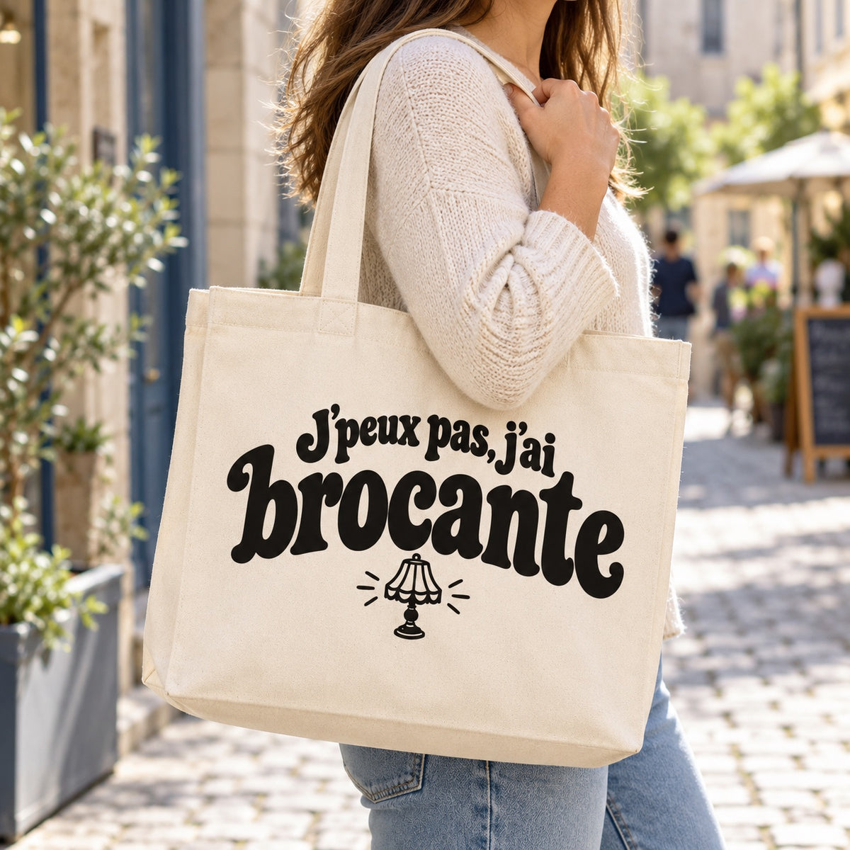 Tote bag « J'peux pas, j'ai brocante » Premium porté à l'épaule dans une rue piétonne ensoleillée
