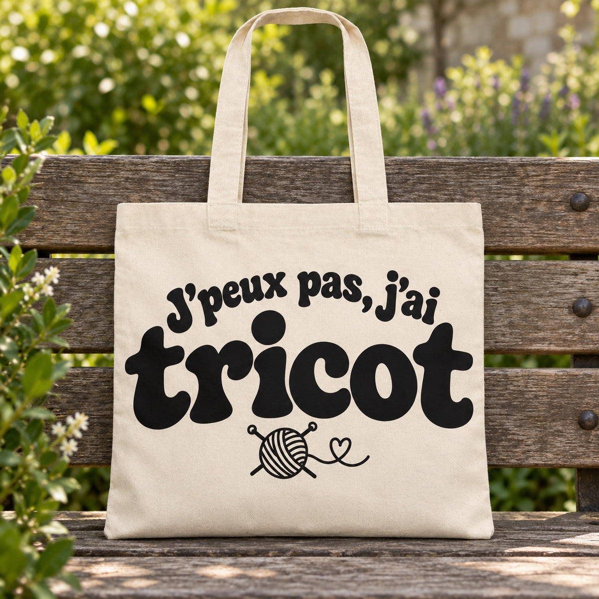 Tote bag « J'peux pas, j'ai tricot » Essentiel posé sur un banc en bois dans un jardin