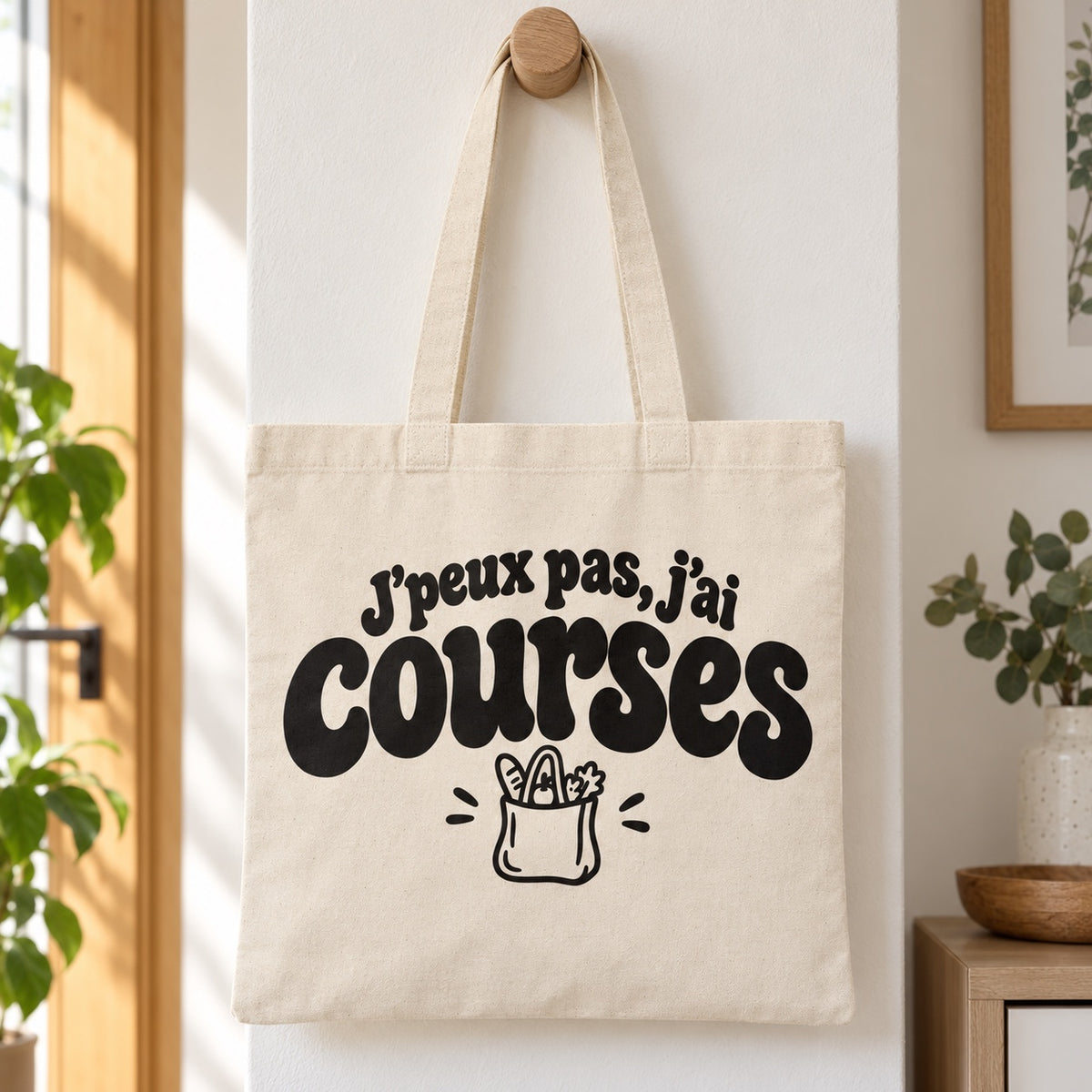 Tote bag « J'peux pas, j'ai courses » Essentiel suspendu à une patère dans une entrée lumineuse