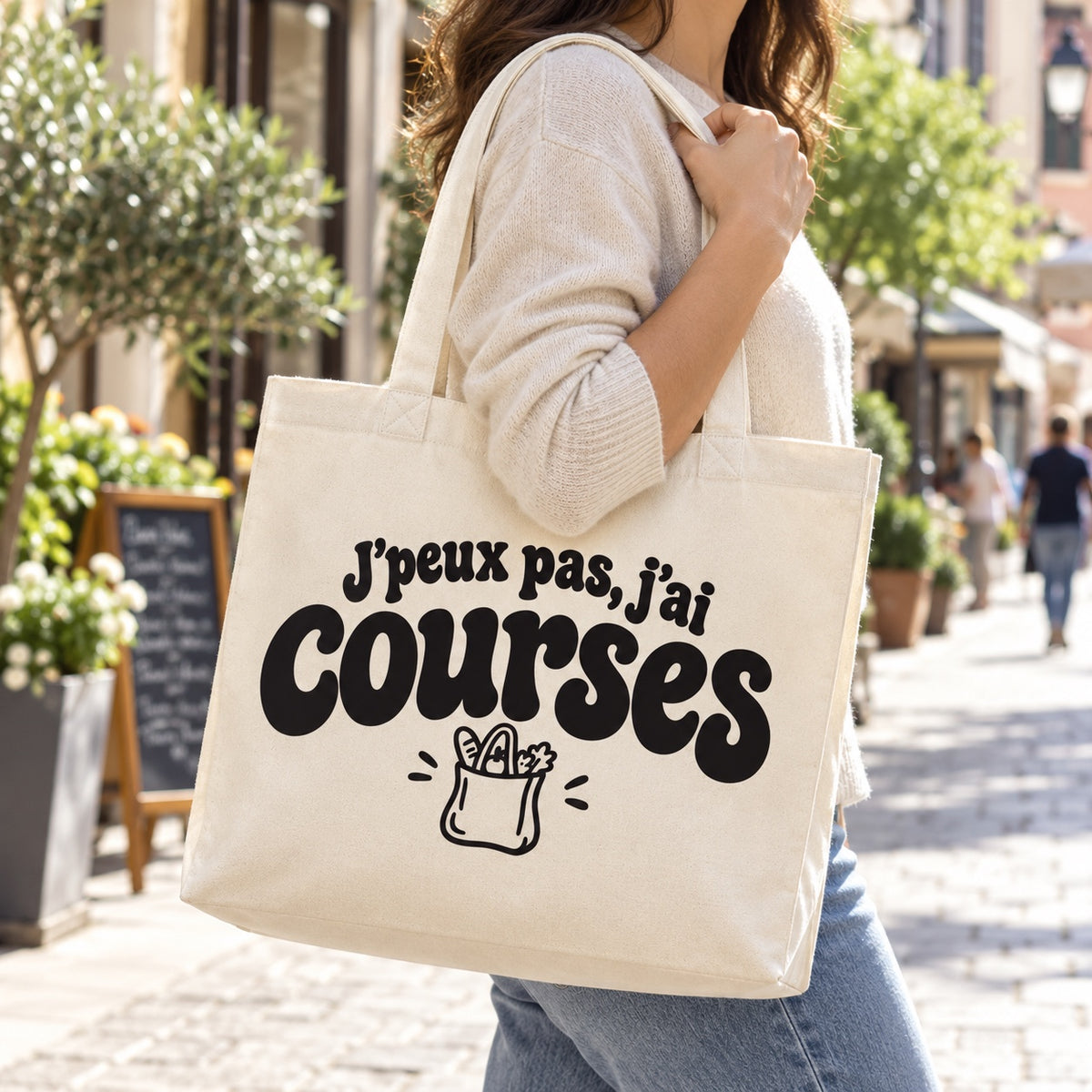 Tote bag « J'peux pas, j'ai courses » Premium porté à l'épaule dans une rue piétonne ensoleillée