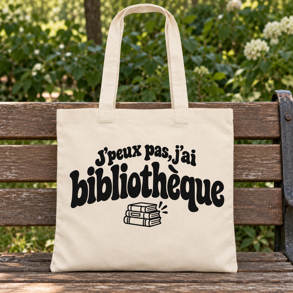 Tote bag « J'peux pas, j'ai bibliothèque » Essentiel posé sur un banc en bois dans un jardin