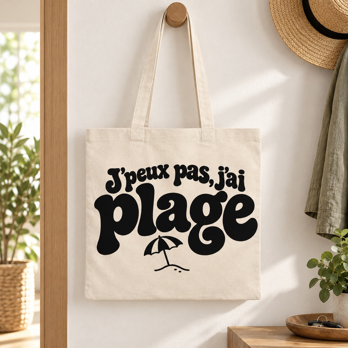 Tote bag « J'peux pas, j'ai plage » Essentiel suspendu à une patère dans une entrée lumineuse